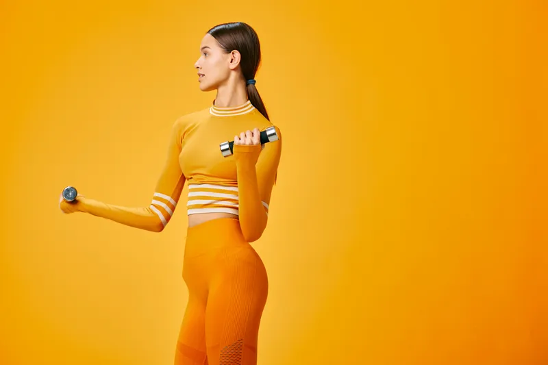 Woman fitness, sport: a fit woman in an orange fitness outfit lifting dumbbells in studio, side profile pose showing toned muscles and confident posture against a solid warm yellow background.