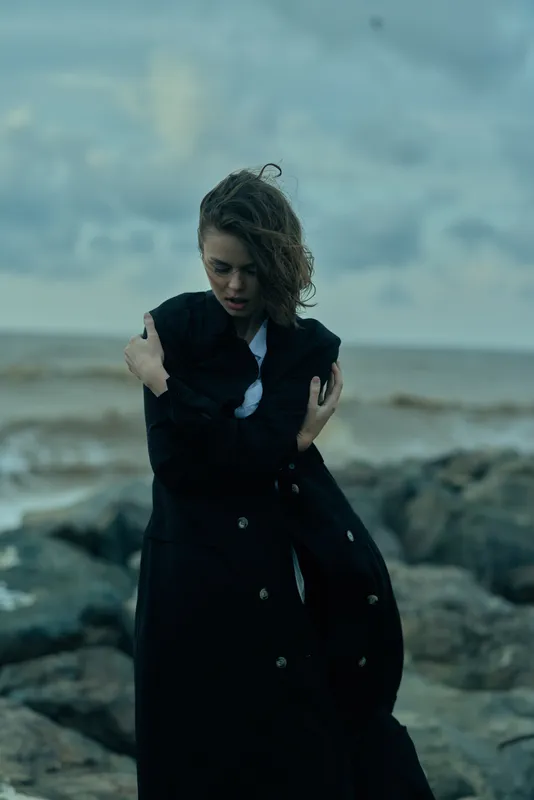 Woman in a dark coat shields herself on a rocky shoreline, wind and moody sea create dramatic outdoor fashion moment, solitary figure amid stormy weather and rugged coastal landscape