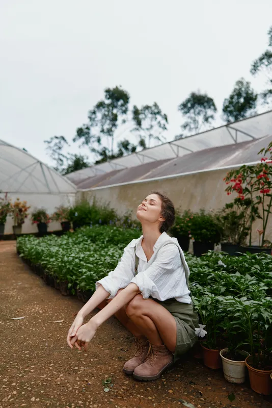 Young woman in a greenhouse, enjoying nature, wearing a light shirt and shorts, surrounded by lush plants, exuding a sense of tranquility and connection to the environment