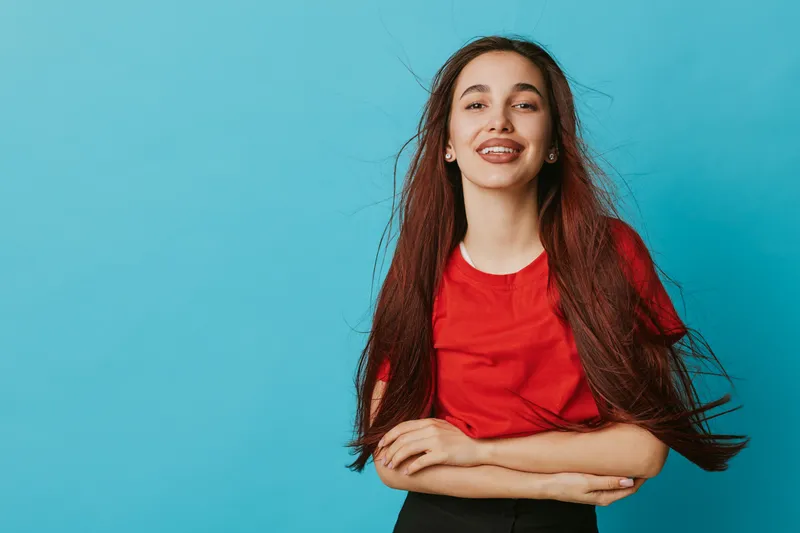 Young woman with long hair wearing red t shirt smiling with crossed arms on blue background. Casual portrait of happy female with natural look and confident pose in studio.