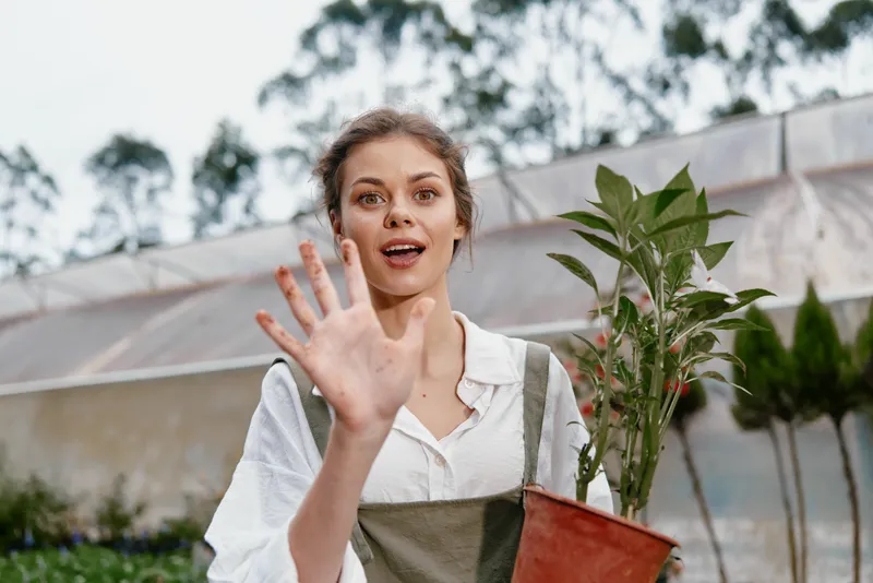 Young woman in a greenhouse, smiling and waving, holding a potted plant, representing joy and connection to nature