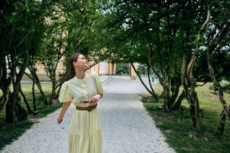 Woman in yellow dress walking on gravel park path under tree canopy, portrait looking up with serene expression, natural light, outdoor fashion and authentic inclusivity