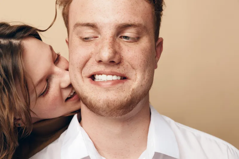 Young couple in casual white shirts share intimate moment, woman kissing man on cheek. Beige background, natural light, concept of love, affection, and close relationship.