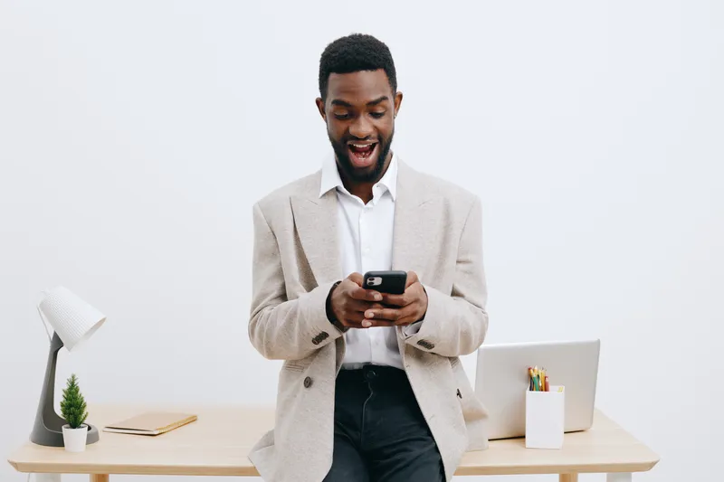 Smiling man using smartphone in modern workspace, casual business attire, happy expression, standing near desk with laptop, papers, and plant, bright minimalist interior, professional and cheerful moo