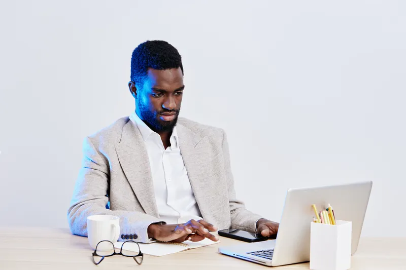 Professional young african-american man working on laptop at modern workspace, wearing stylish light blazer and white shirt, focused on screen, office accessories on desk, minimalistic background, bus