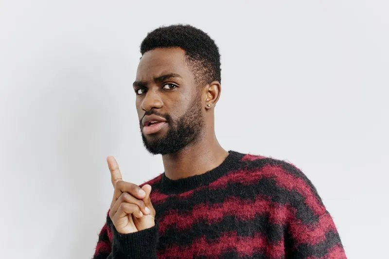 Уверенный мужчина в красно-чёрном свитере указывает прямо на камеру с решительным выражением лица, передавая ощущение силы