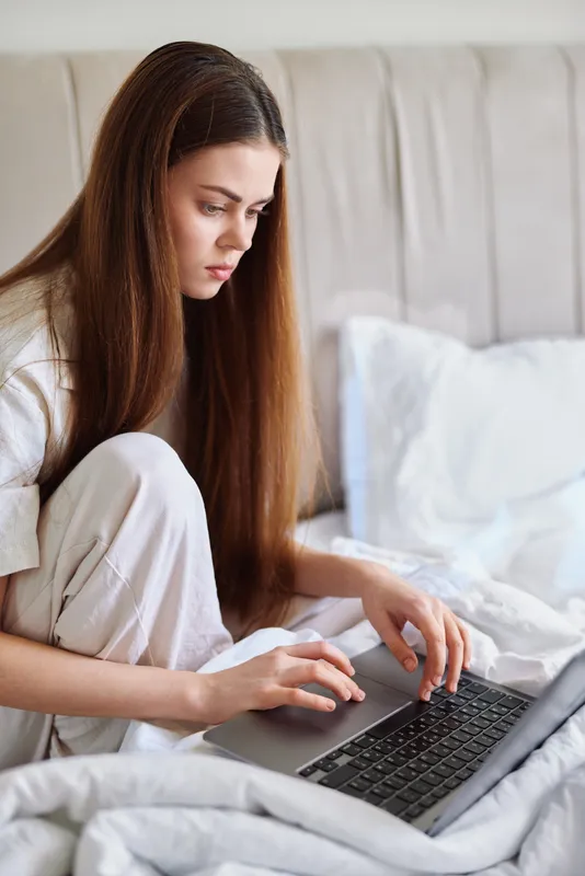 Woman teenager on bed in pajamas using laptop in bedroom, focused on screen and typing, remote study or work from home with cozy blankets and natural light.
