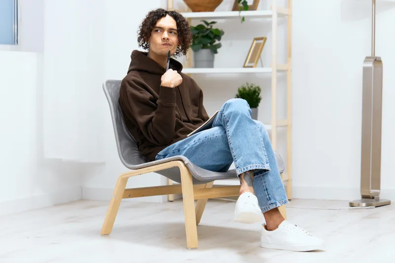 Парень с вьющимися волосами сидит на стуле с блокнотом и ручкой на светлом фоне