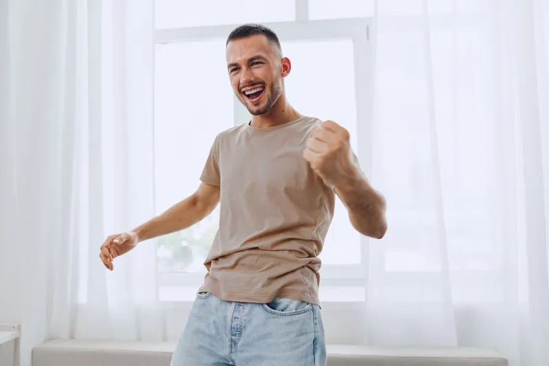 Joyful man celebrating success indoors with bright sunlight pouring through the window. His energetic pose captures a sense of happiness and positivity in a modern space.