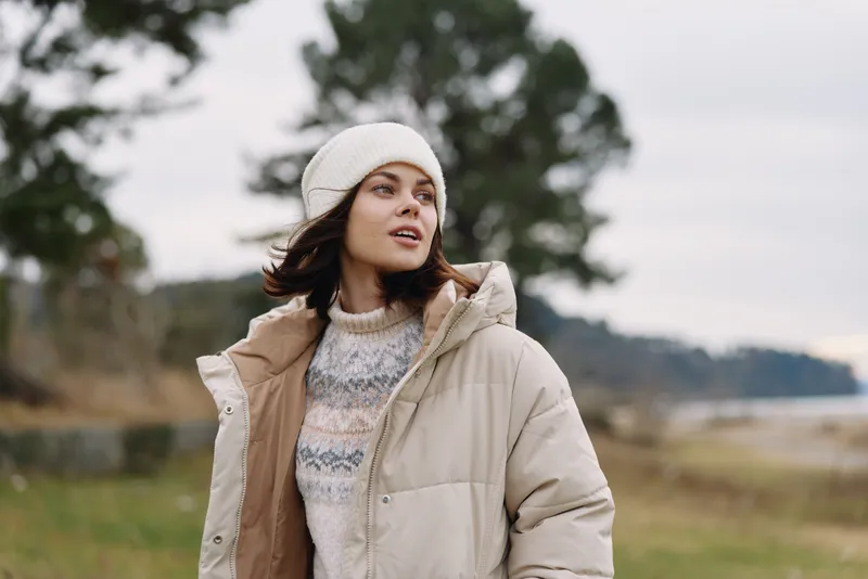 Woman in a warm winter jacket and knit sweater standing outdoors in an open field, wearing a beanie, gazing upward with soft natural light and distant trees.
