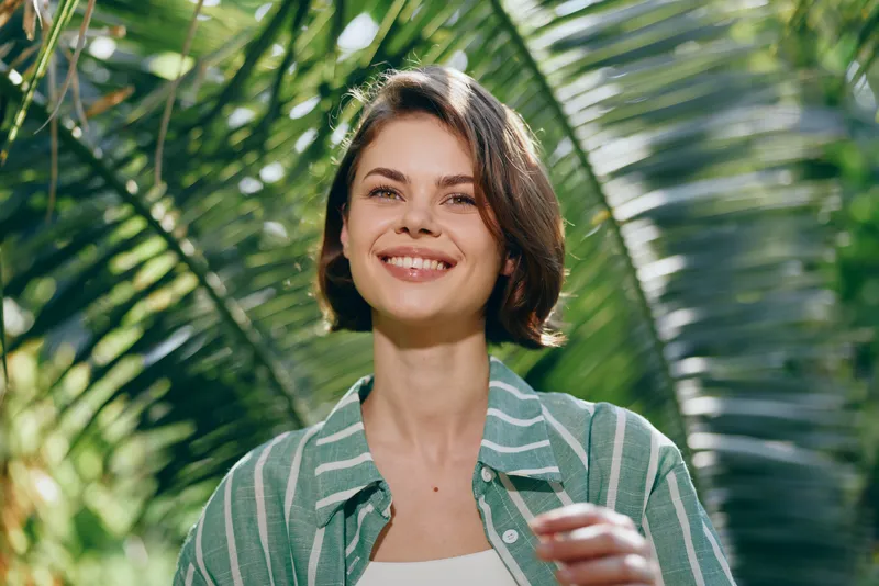 Smiling woman stands outdoors amid lush tropical greenery, wearing a striped green shirt, capturing a bright, relaxed summer vibe.