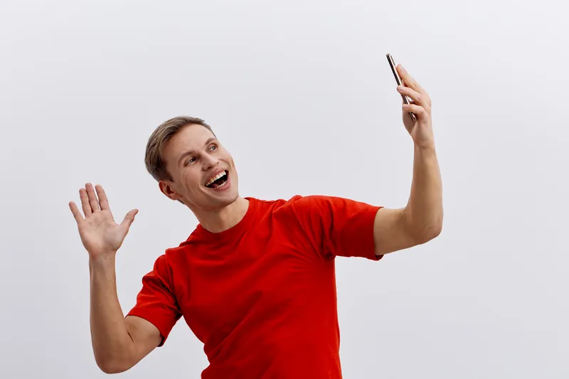 Smiling young caucasian man wear red t-shirt hold smartphone in hand wave gesture video call on plain light background digital nomad concept