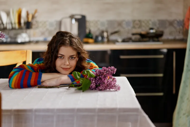 Woman relaxing indoors at a kitchen table, resting her chin on crossed arms, lilac bouquet nearby, warm natural light, casual home moment, soft colors, cozy atmosphere, blissful, serene