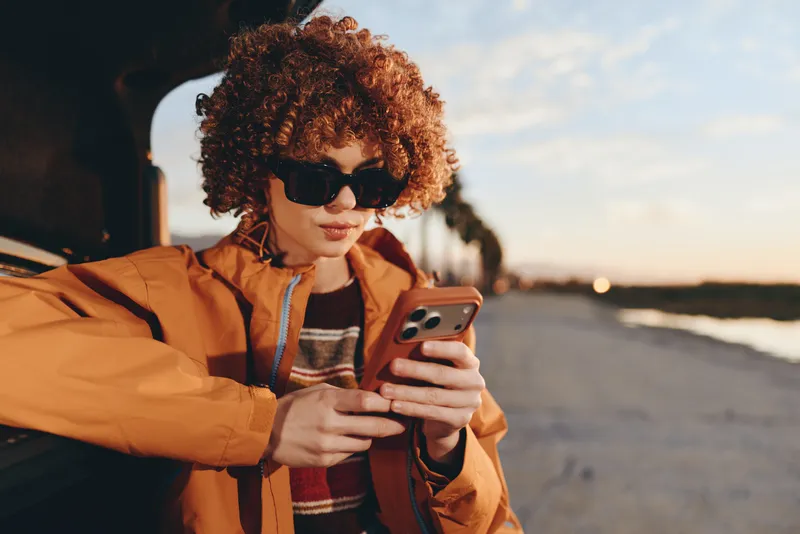 Woman lifestyle outdoors, curly hair and sunglasses, holding smartphone by car, warm sunset glow, casual jacket, relaxed travel mood, coastal road background, soft wind, evening sky