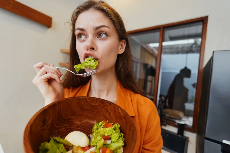 Молодая женщина в повседневной одежде откусывает кусочек свежего салата в теплой кухне, что отражает здоровый образ жизни и пользу питательной еды для хорошего самочувствия