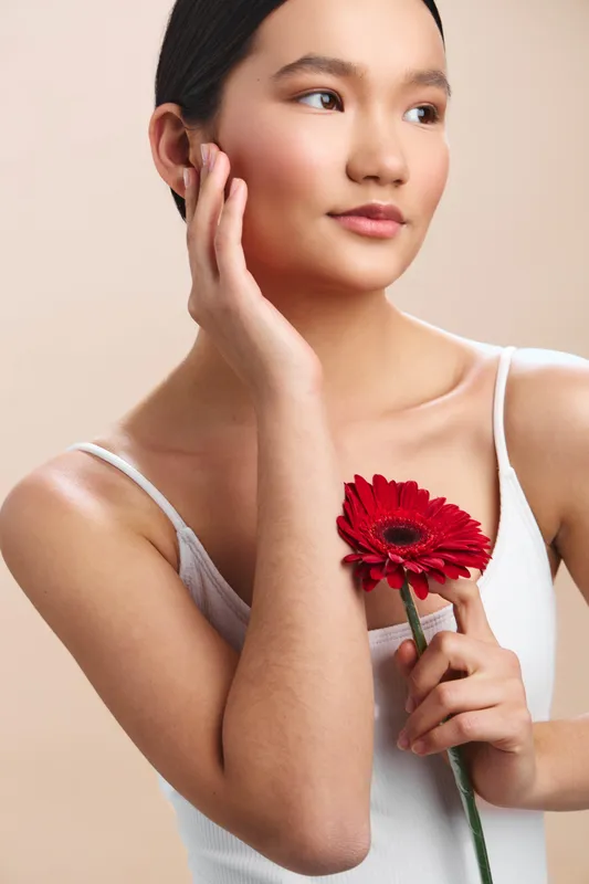 asian woman holds a vibrant red flower near her shoulder, her calm gaze distant yet warm, while soft studio lighting flatters her smooth skin, creating a serene, intimate portrait with a gentle,