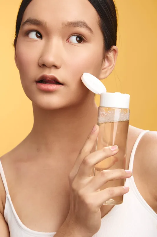 Asian woman applies skincare product with a cotton pad on her face in a bright studio setting, highlighting a fresh beauty routine and confident selfcare in a calm, modern atmosphere.