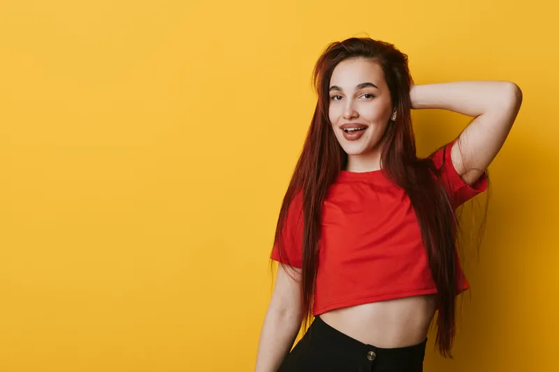 Young woman with long hair wearing red crop top posing with hand behind head smiling and sticking out tongue against yellow background in studio portrait.
