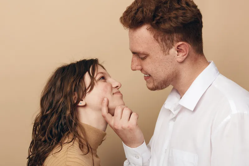 Young couple in casual clothes share a tender moment, man gently touching woman’s cheek. Warm beige background creates intimate and loving atmosphere, concept of romance and affection.