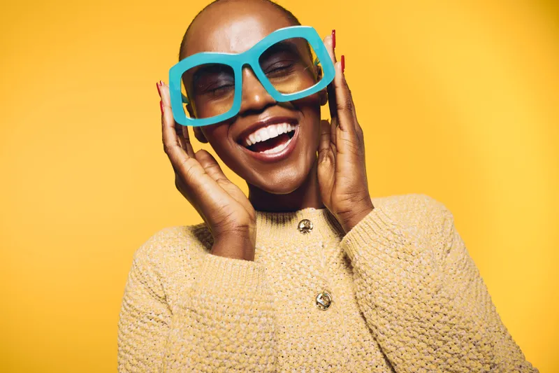 Happy smiling black woman with dark skin wearing oversized blue glasses and beige sweater stands against yellow background. Concept of joy, fashion, and positive emotions in studio.