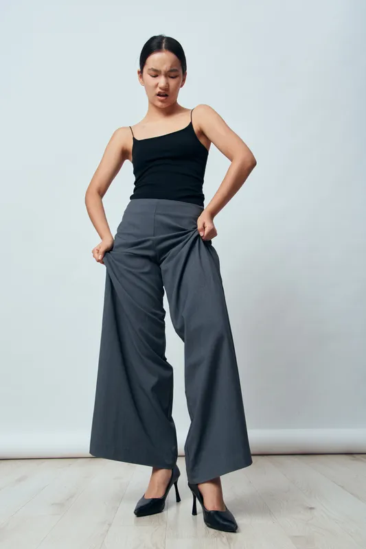 Young woman wearing black tank top and wide leg gray pants stands with hands in pockets and an expressive face on light background in studio. Fashion, casual, modern style look.
