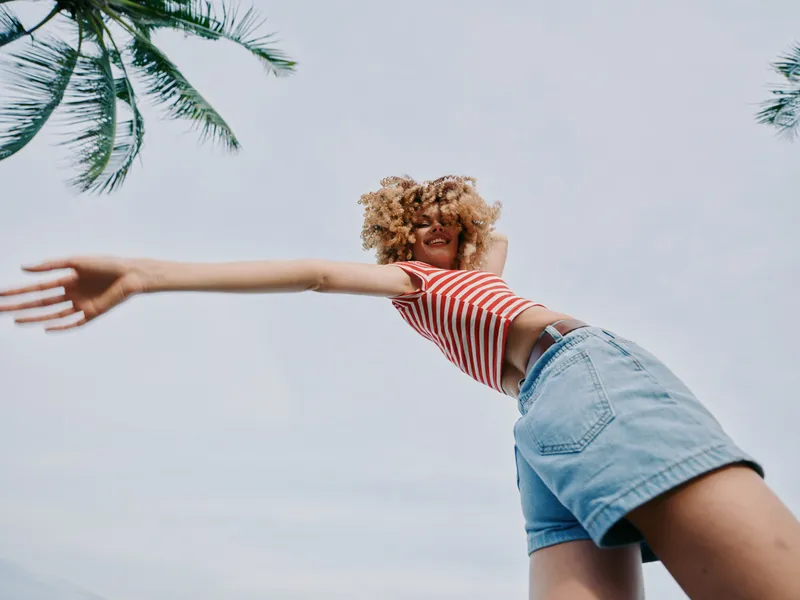 A carefree young woman dances outdoors under a bright sky, wearing a red striped crop top and denim shorts, arms extended in a joyful, sunlit pose.