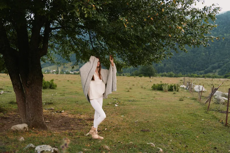 Cheerful woman outdoors, relaxed pose under a leafy tree, white outfit, sunlit meadow background, gentle breeze, natural colors, peaceful countryside vibe, lifestyle moment