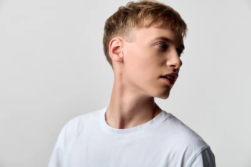 Young man in a white tee turns his head to the side, presenting a calm studio vibe with a clean background, natural lighting and minimal wardrobe for a modern casual look.