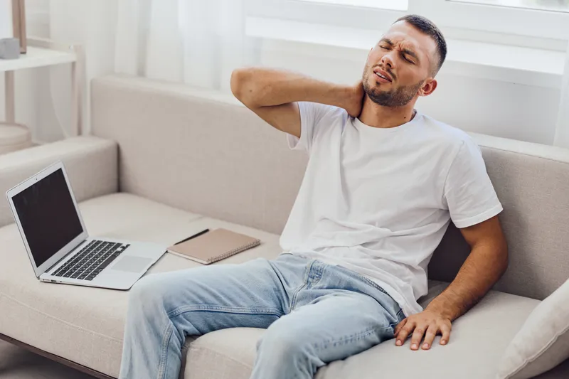 Relaxed man experiencing neck pain while sitting on a sofa with a laptop, showcasing the importance of ergonomic workspace design, and health awareness in modern life.