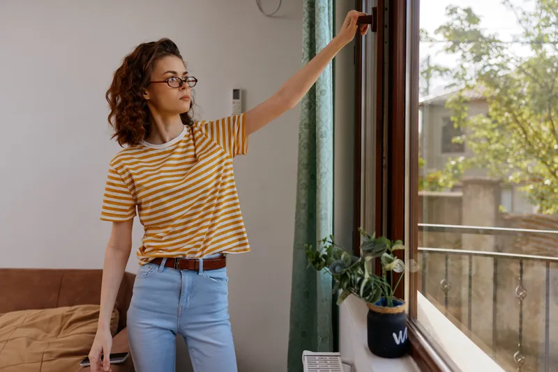 A woman with glasses in a striped shirt and jeans stands indoors, opening a window to welcome daylight. A casual lifestyle moment inside a warm home interior.