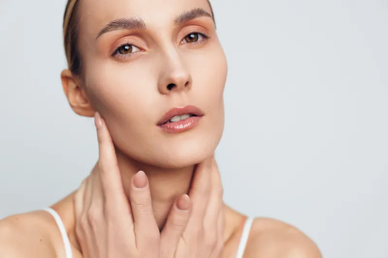 A close up portrait of a woman with clear skin, soft makeup, and a hand resting near her jaw, conveying calm beauty and skincare.