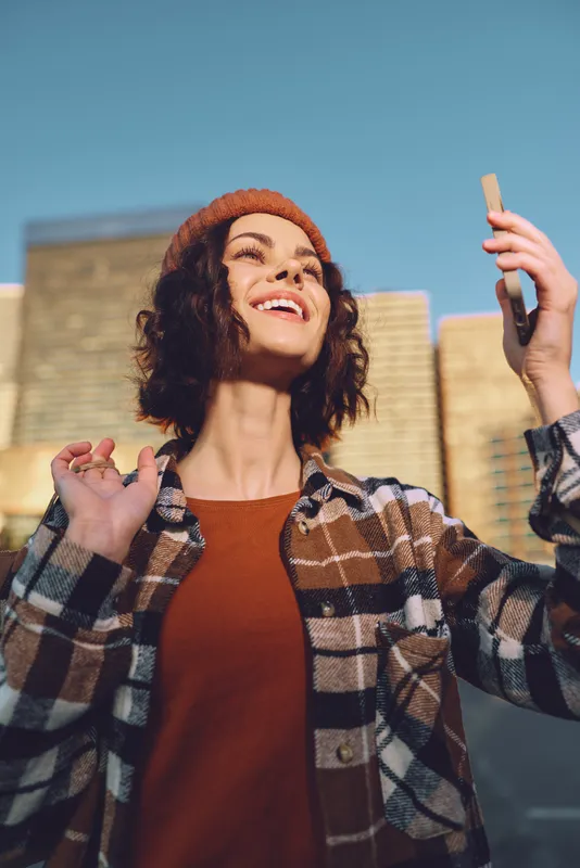 woman smile phone urban candid golden hour beanie authenticity mindful living emotional storytelling. Young woman grins holding phone for a warm golden hour glow selfie, candid lifestyle portrait in