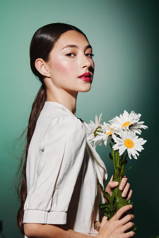 Young woman with sleek glossy hair and red lipstick holds daisies, wearing a white blouse against green background. Creative beauty editorial with natural elegance.