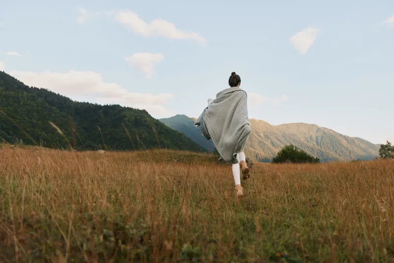 Woman running through a sunlit meadow with a flowing scarf, embracing outdoor lifestyle and freedom in a mountainous landscape, motion and fresh air captured.
