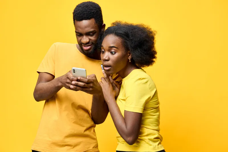 Happy young couple enjoying moment together, looking at smartphone with excitement against vibrant yellow background, casual outfits exuding playful energy