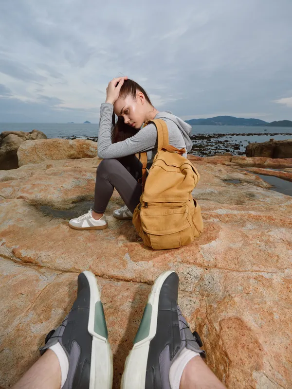 Young woman in sporty outfit with backpack sitting on rocky shore, gazing into the distance, capturing a moment of contemplation by the seaside