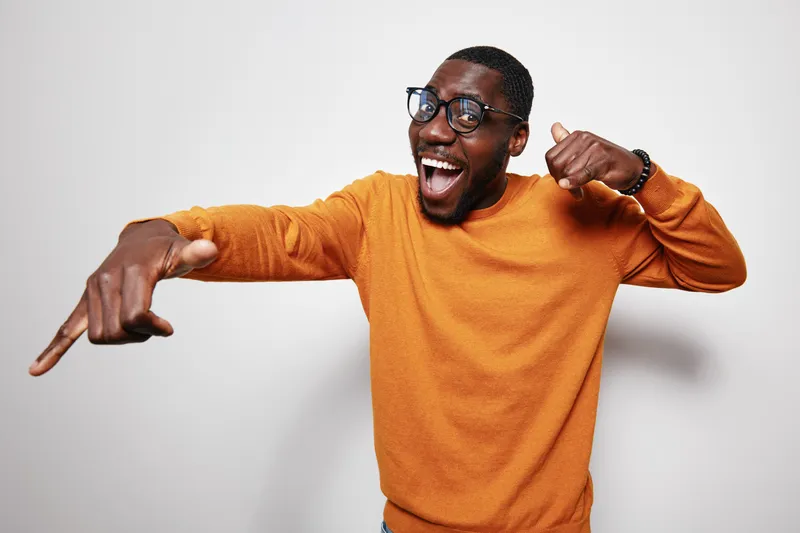 African American man with dark skin wearing glasses and orange sweater pointing with excitement and smiling broadly against white background in studio portrait.