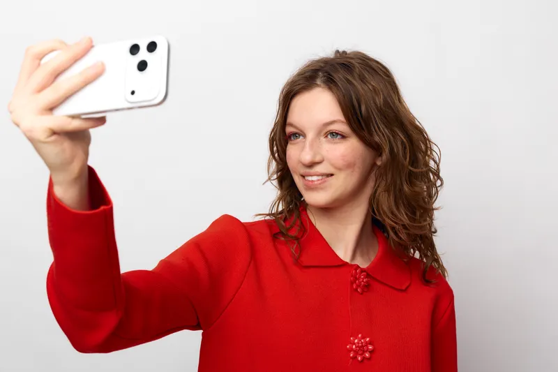 Young woman with curly hair wearing red sweater takes selfie with smartphone on white background. Concept of social media, technology, communication, and casual lifestyle.