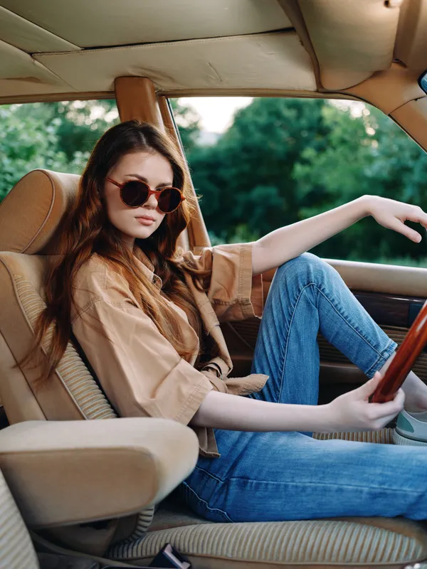 Young woman in sunglasses sitting in a vintage car, wearing a casual outfit, showcasing a relaxed summer vibe with greenery in the background.
