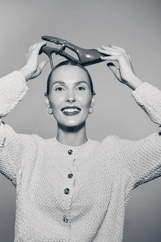 Smiling caucasian woman with fair skin holding a high heel shoe above her head. Studio portrait in black and white. Concept of fashion, style, and happiness.