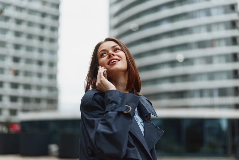 Confident businesswoman speaks on her phone outside in a modern city, wearing a dark trenchcoat and looking up with a bright smile amid glass towers.