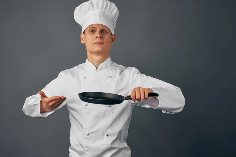 Красивый мужчина в поварской форме держит сковороду в руках на кухне, занимаясь приготовлением пищи