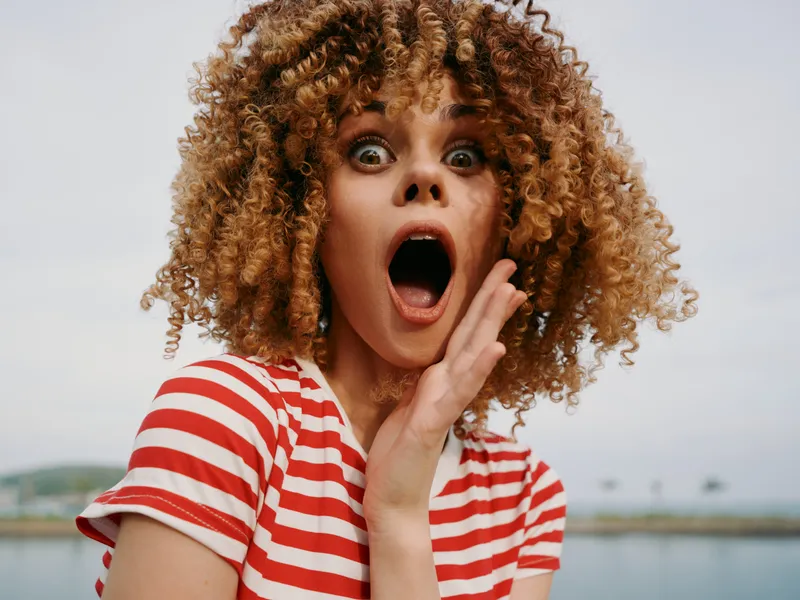 Surprised young woman with voluminous curly hair and wide eyes wearing a red striped shirt outdoors near water, capturing dramatic astonishment and candid emotion in bright color.