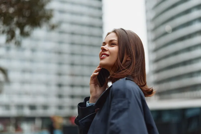 Smiling woman on a city street talks on a cellphone, wearing a dark jacket, blurred skyscrapers in the background, urban portrait captured outdoors with natural light
