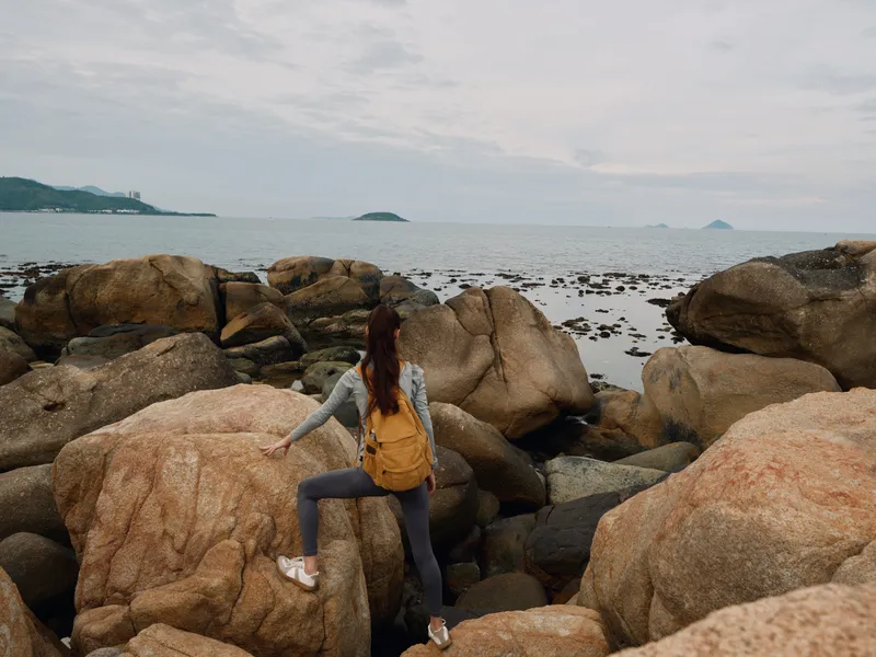 Young woman exploring rocky coastline, wearing activewear with a backpack, facing the sea, evoking a sense of adventure and nature s beauty