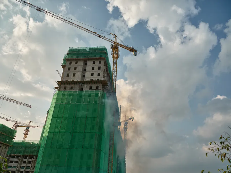 Высокое здание, находящееся в процессе строительства, покрыто зелёной сеткой, на фоне пушистых облаков и голубого неба работают краны