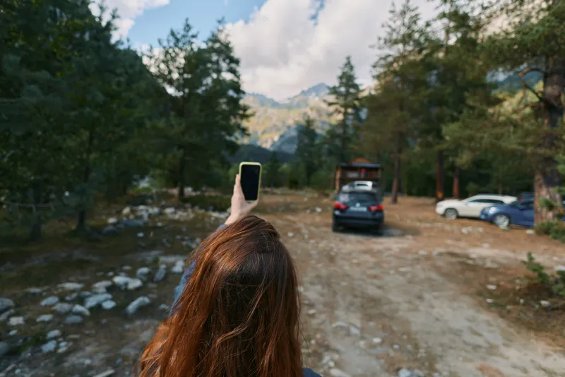 A person with long hair is seen from behind holding up a smartphone to take a photo in a forested parking area. Cars and distant mountains frame a bright outdoor scene.