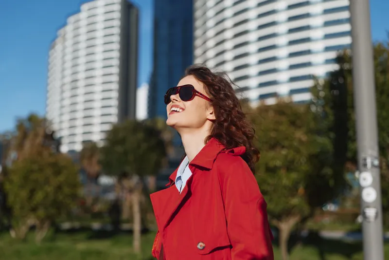 Smiling woman in a red coat looks up at the sunny sky in a city park with modern buildings behind, conveying confidence and warmth in a stylish urban moment.