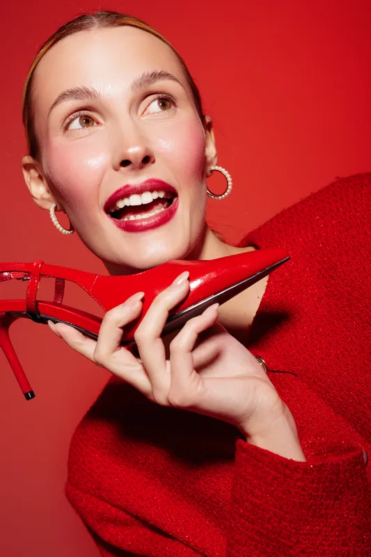 Smiling woman in red sweater holds red high heel shoe near face on red background. Concept of fashion, style, beauty, and vibrant color contrast in studio portrait.