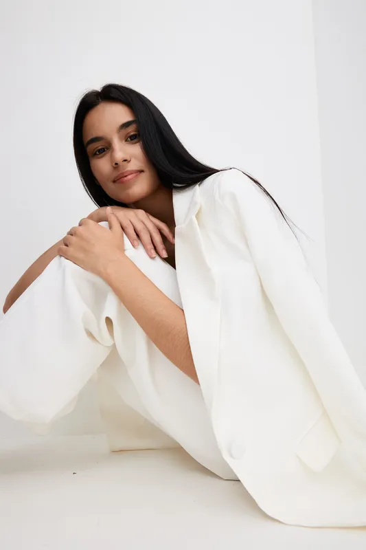 young woman in white suit sitting on floor, with long dark hair and relaxed expression against minimalistic white background, casual modern style